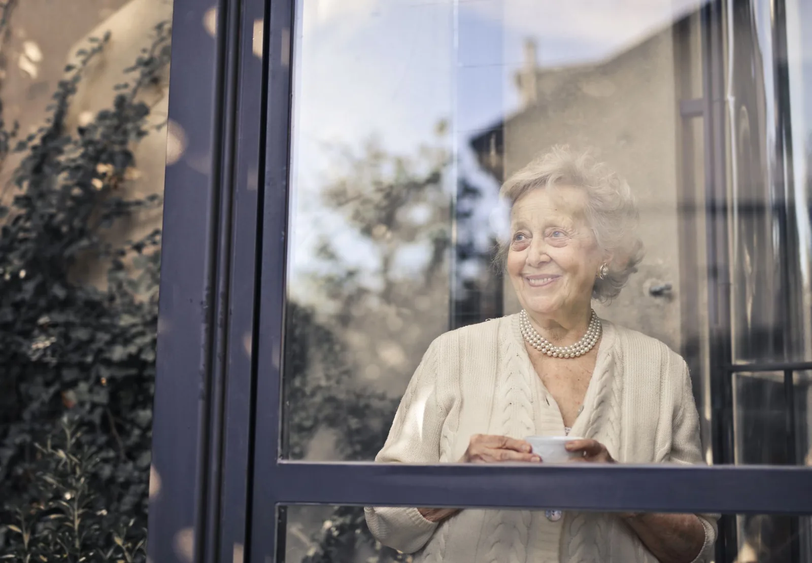 An older woman with a warm smile holds a cup of tea by a sunlit window at home.